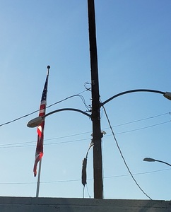 A limp US flag hangs against a flat blue sky, with industrial power & light poles in the foreground. 